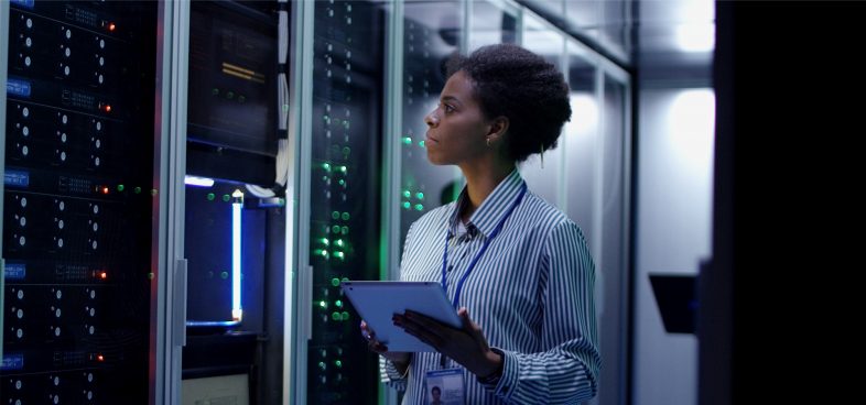 A Black woman stands in a dimly lit server room, holding a tablet and examining network equipment, illuminated by the blinking blue and green lights of the servers.